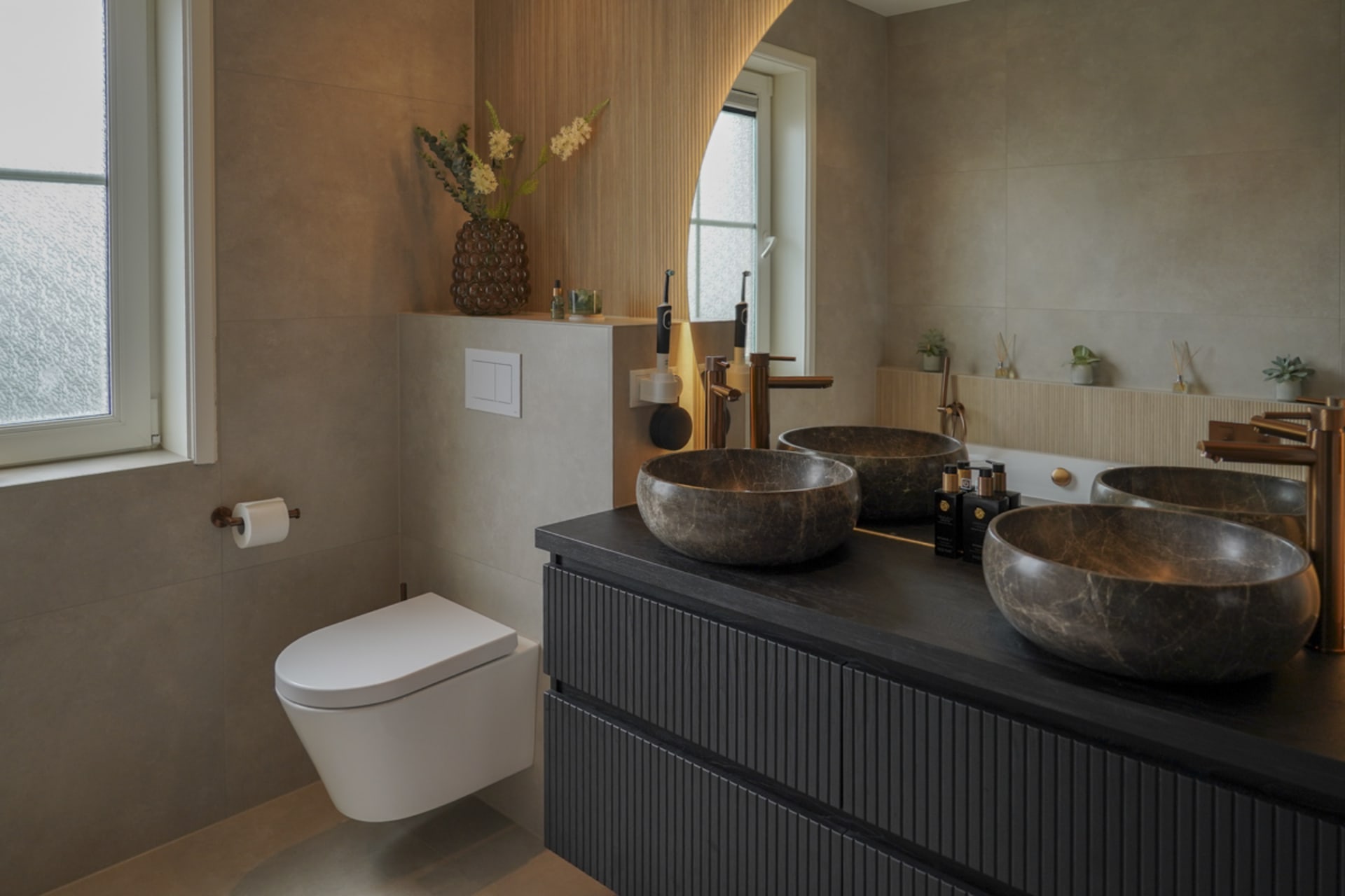 Overzicht van de badkamer met beige vloertegels, zwart ribbelmeubel, ronde spiegel en hangend toilet