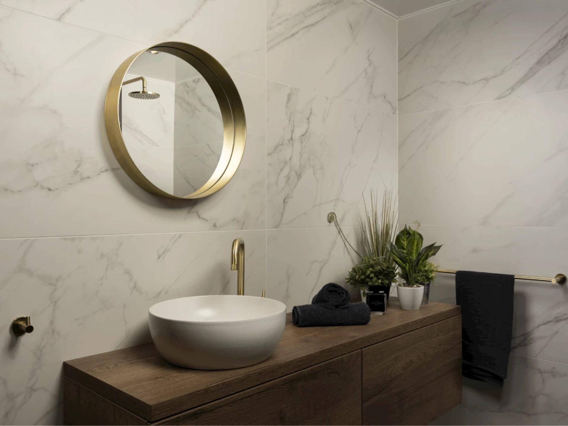 Lichte marmerlook wandtegels met grijze adering, met een ronde spiegel met gouden omlijsting boven het houtlook badmeubel