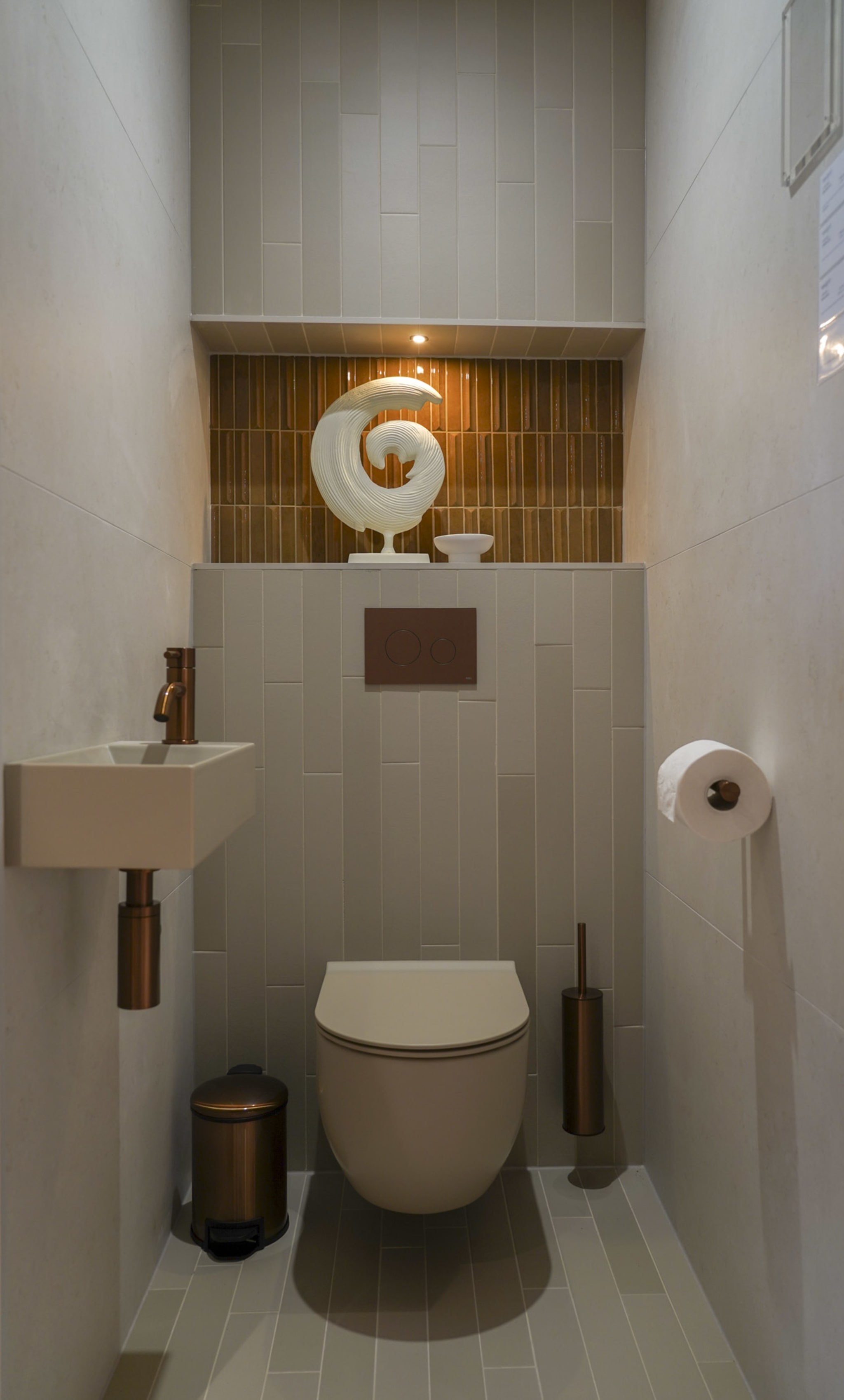 Een compacte beige toiletruimte met verticale tegels, een hangtoilet, een klein fonteintje en koperkleurige accenten.