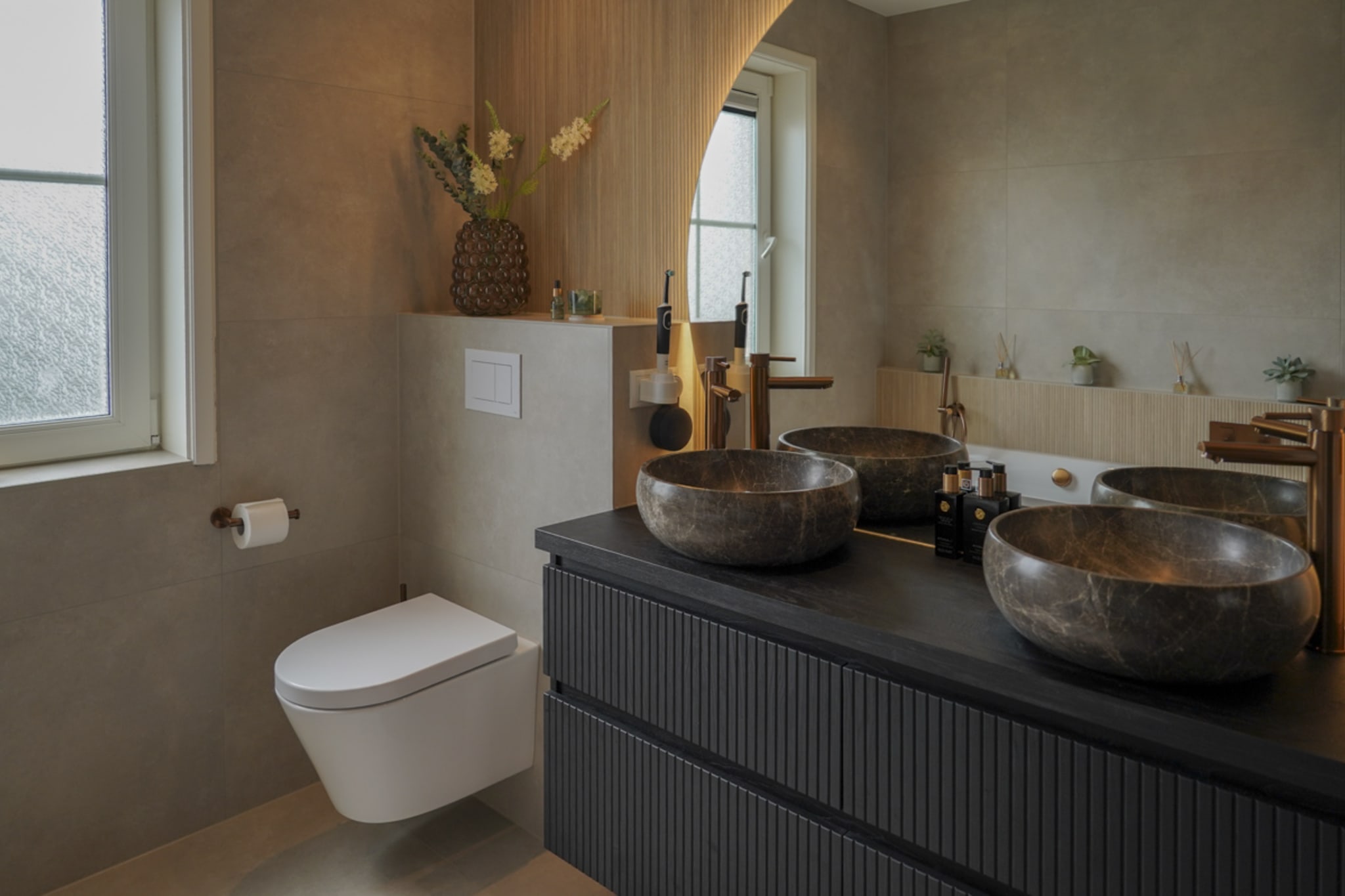 Overzicht van de badkamer met beige vloertegels, zwart ribbelmeubel, ronde spiegel en hangend toilet