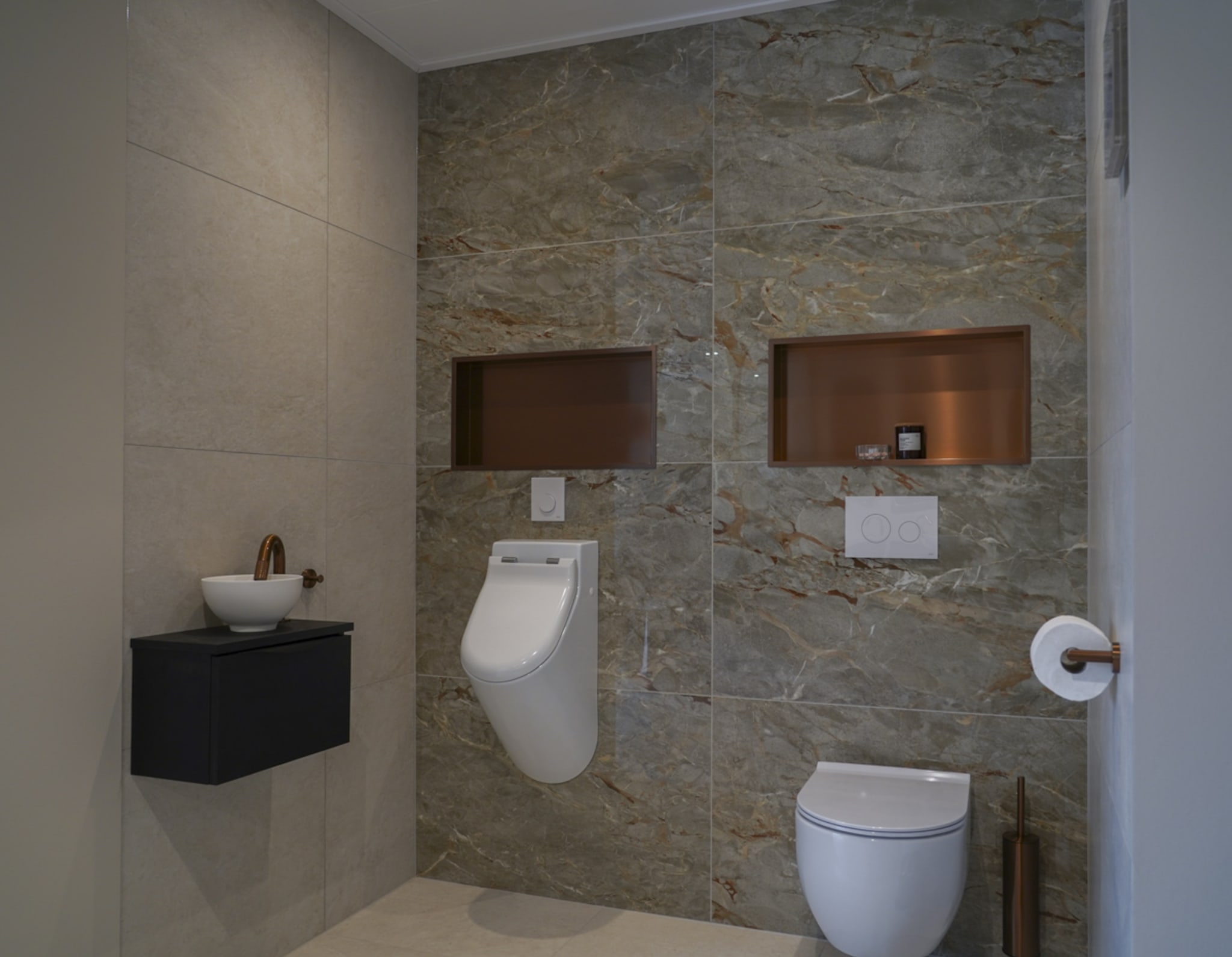 Moderne toiletruimte met wandcloset, urinoir, fontein en koperkleurige accenten op marmerlook wandtegels.