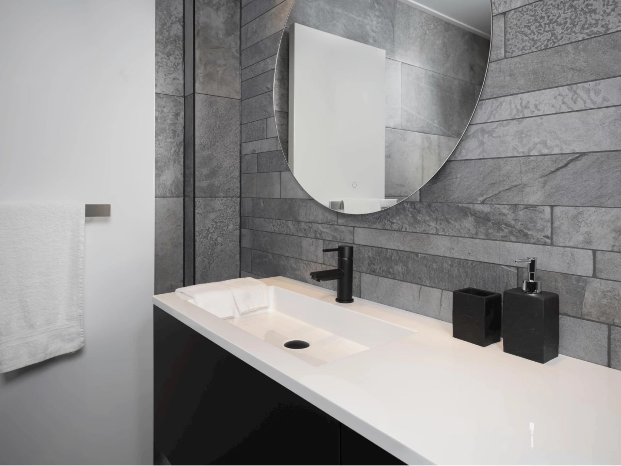 Zwart badmeubel met een dunne witte wastafel van solid surface, mat zwarte kraan en een ronde spiegel tegen de grijze strooktegels