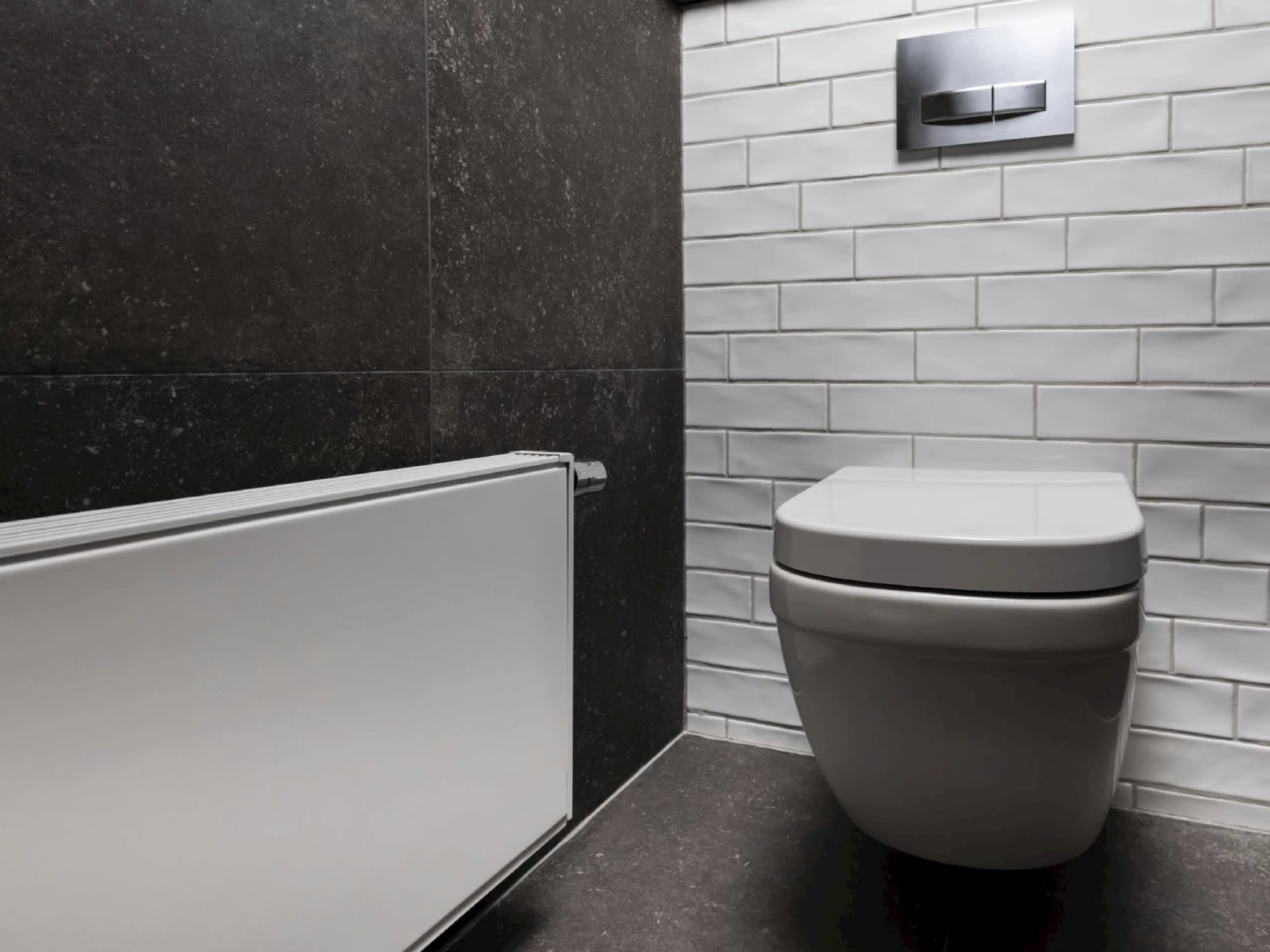 Witte radiator voor de donkere terrazzolook wand, met rechts het wandcloset tegen de witte tegels