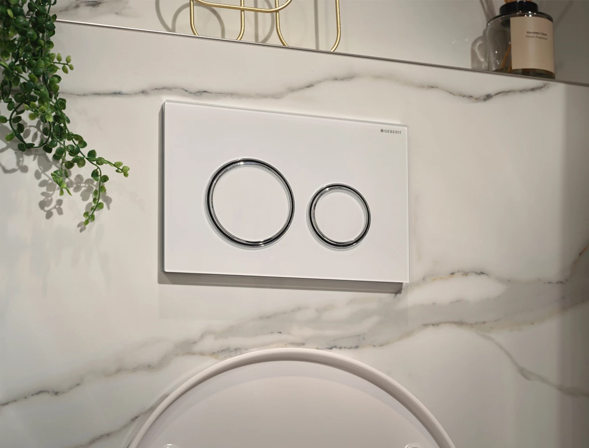 Witte drukplaat van Geberit met twee verchroomde ringen op de marmerlook wand, met een richel erboven