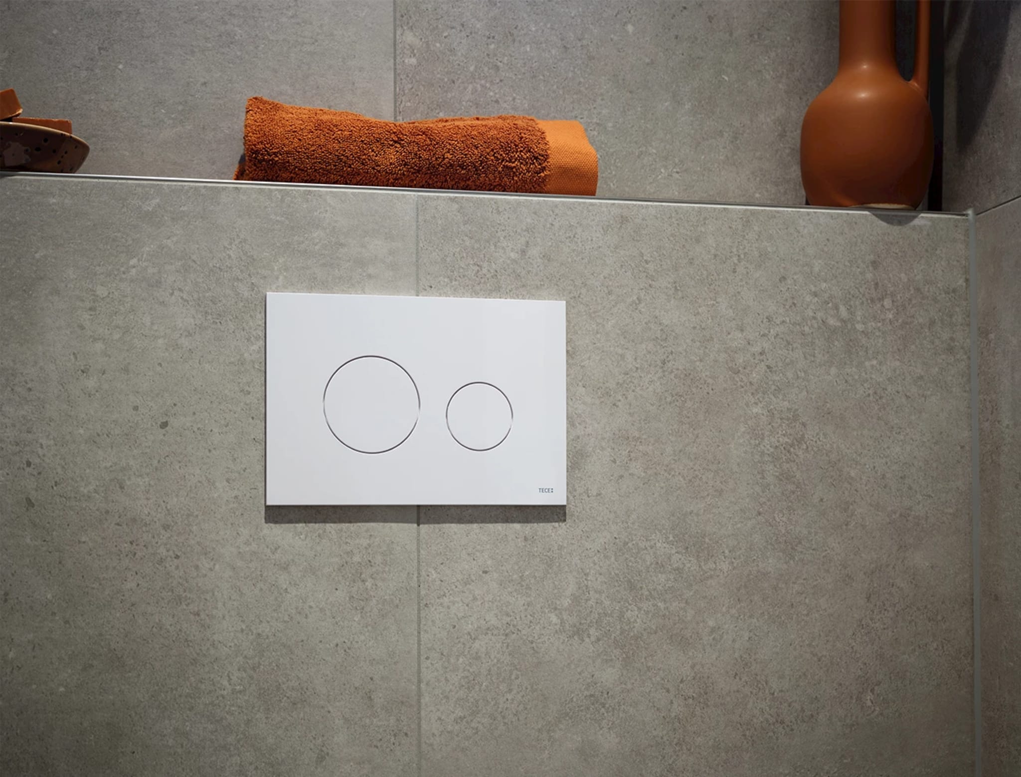 Witte drukplaat van TECE met twee ronde knoppen op de grijsbeige wand, met daarboven het plateau met een handdoek en een vaas