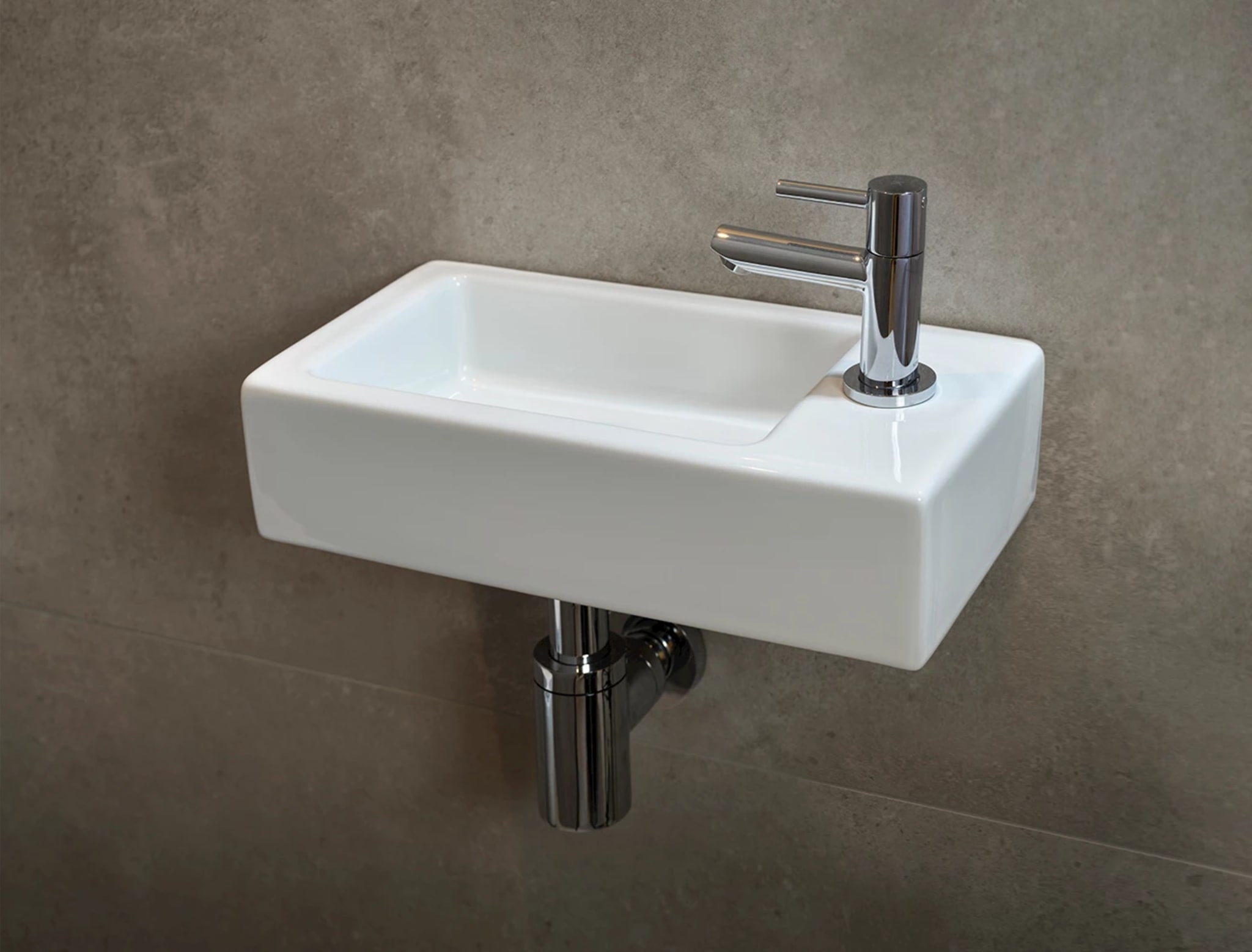 Klein wit keramisch fonteintje met een chromen eenhendelkraan en chromen sifon tegen de grijsbeige betonlook wand