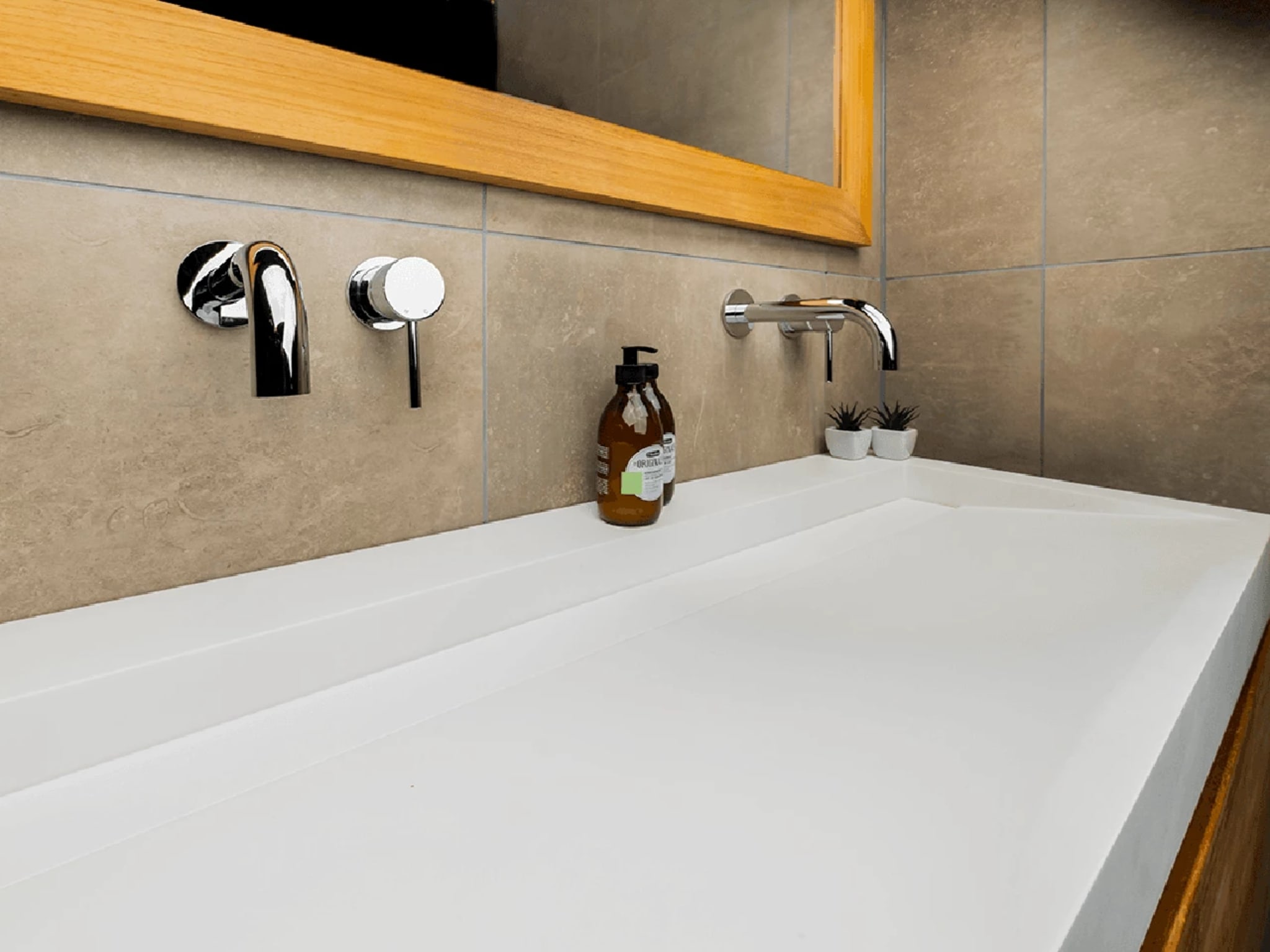 Detail van de mat witte wastafel van solid surface met twee chromen inbouwkranen onder de teakhouten spiegellijst