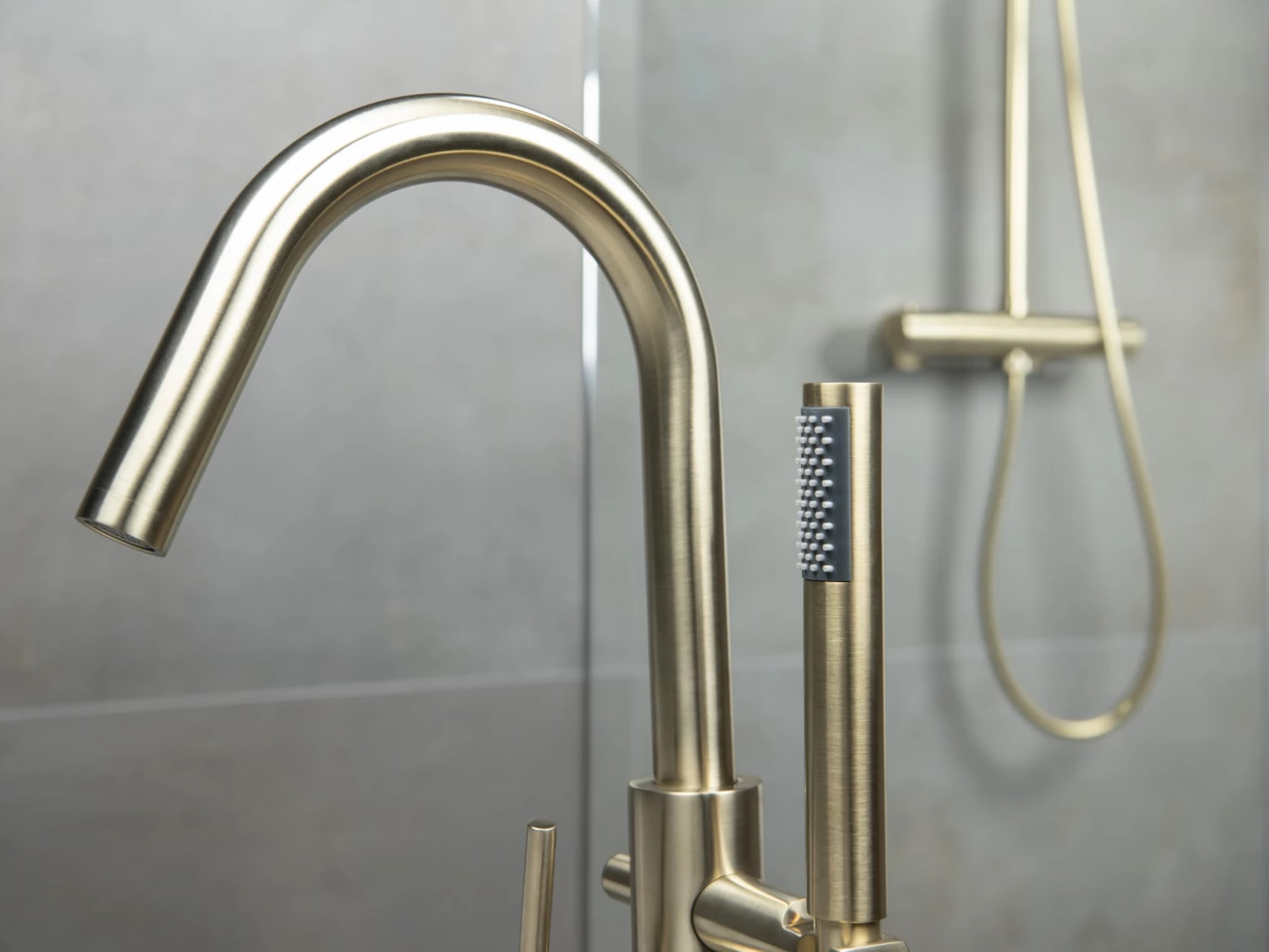 Detail van de vrijstaande badkraan met slanke handdouche, met achter het glas de gouden regendoucheset