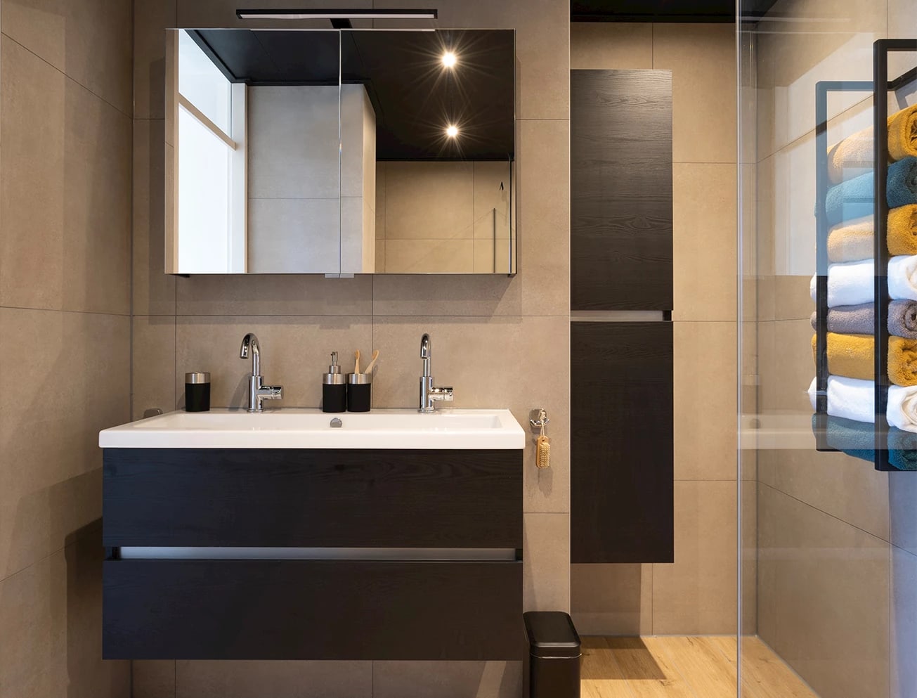 Kranen industriële badkamer: donker wastafelmeubel met witte wasbak, twee chromen kranen en een groot verlicht spiegelkastje tegen beige tegels